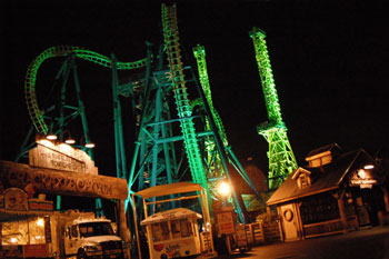 night with lit carnival booths and food stands in front of an illuminated Goliath Coaster Thrill Ride with vibrant colors of yellow, blue and purple by Studio Due CityColor 2000 architecural lighting fixture, Six Flags New England - Agawam, Massachusetts, USA